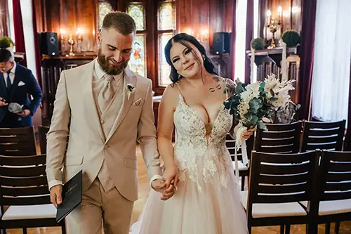 Ein Paar in Hochzeitskleidung schreitet anmutig den Gang entlang. Die Braut, einen Blumenstrauß in der Hand, beeindruckt in ihrem tief ausgeschnittenen Kleid. Der Bräutigam im hellen Anzug hält eine Broschüre in der Hand. Holzstühle und Buntglas bilden die Kulisse, wunderschön eingefangen von Hochzeit in Tharandt-Melanie Kunert Fotografie.