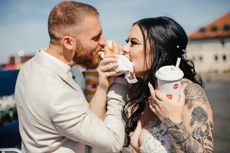 Braut und Bräutigam in Hochzeitskleidung genießen während ihrer Hochzeit einen Burger und Limonade. Der Bräutigam trägt einen hellen Anzug, während die tätowierte Braut in ihrem weißen Kleid strahlt. Ihre Lächeln strahlen und strahlen pure Freude vor der bezaubernden Kulisse aus.