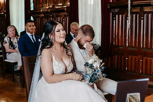 In einem holzgetäfelten Raum strahlt eine Braut im weißen Kleid neben ihrem Bräutigam, der mit Tränen in den Augen im hellen Anzug ihren Brautstrauß umklammert. Melanie Kunert Fotografie hält diesen besonderen Moment während ihrer Hochzeit in Tharandt fest, während die Gäste gemütlich hinter ihnen sitzen.