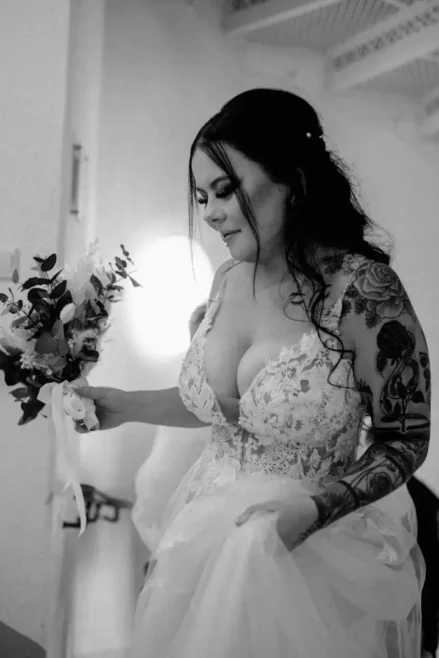 In einer zeitlosen schwarz-weißen Kulisse hält die Braut in einem detailreichen Spitzenbrautkleid einen Blumenstrauß. Ihr heiterer Gesichtsausdruck spiegelt die Romantik der Hochzeit in Tharandt wider. Ihr Haar fällt in Wellen herab, während Tattoos auf ihrem Arm der eleganten Szene eine einzigartige Note verleihen.