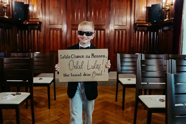 Ein kleiner Junge mit Sonnenbrille und Anzug steht in einem Holzraum und hält ein Schild mit der Aufschrift „Letzte Chance zum Rennen, Onkel Lukas! Hier kommt deine Braut!“. Diese charmante Szene spielt sich bei einer Hochzeit in Tharandt ab, wo auf beiden Seiten des Raumes Stuhlreihen stehen.