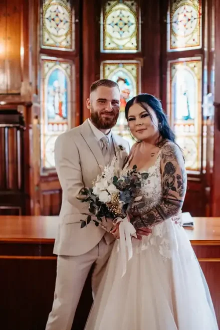 Auf einer bezaubernden Hochzeit in Tharandt, aufgenommen von Melanie Kunert Fotografie, steht ein Paar zusammen in einer Kirche. Die Tattoos der Braut blitzen unter ihrem weißen Kleid hervor, während sie einen Blumenstrauß hält, während der Bräutigam in seinem hellbeigen Anzug ihren Stil unterstreicht. Buntglasfenster tragen zur Schönheit der Szene bei.
