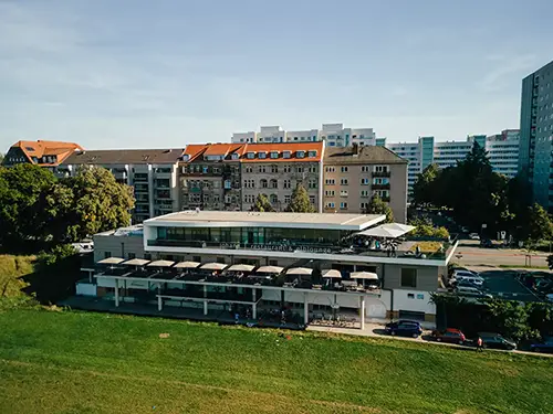 Luftaufnahme eines modernen Restaurants am Flussufer, ideal für eine Hochzeit in Tharandt, mit Außensitzplätzen entlang eines grasbewachsenen Ufers. Dahinter befinden sich historische Gebäude mit roten Dächern und moderne Wohnblöcke. Es ist ein sonniger Tag mit klarem, blauem Himmel.