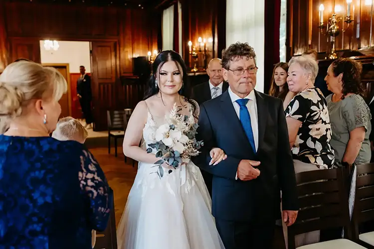 Eine Braut in weißem Kleid und Brautstrauß schreitet Arm in Arm mit einem älteren Mann zum Altar. Gäste beobachten sitzend und stehend, umgeben von Holzwänden und sanftem Licht. Diese wunderschöne Hochzeit in Tharandt wurde von Melanie Kunert Fotografie festgehalten.