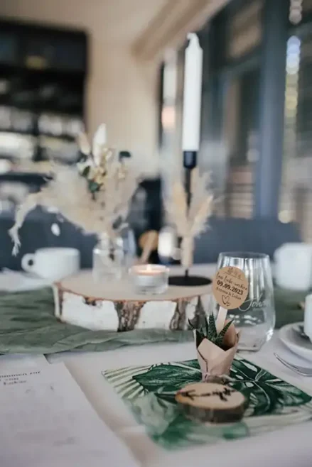 Eine stilvolle Tischdekoration für eine Hochzeit in Tharandt präsentiert sich im Waldthema mit einem hölzernen Kerzenhalter, Pampasgras und einer personalisierten Holzscheibe mit dem Datum. Eine kleine Topfpflanze steht auf einem Tischset mit grünem Blattmuster, dazu eine brennende Kerze und ein Glas.