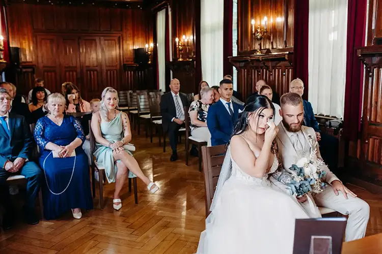 Braut und Bräutigam sitzen in einem dekorierten Raum bei ihrer Hochzeit in Tharandt, umgeben von Gästen in festlicher Kleidung. Wunderschön eingefangen von Melanie Kunert Fotografie, wischt sich die Braut die Augen, während der Bräutigam geradeaus blickt. Der Raum ist mit Holzvertäfelungen, Kronleuchtern und großen Fenstern mit Vorhängen ausgestattet.