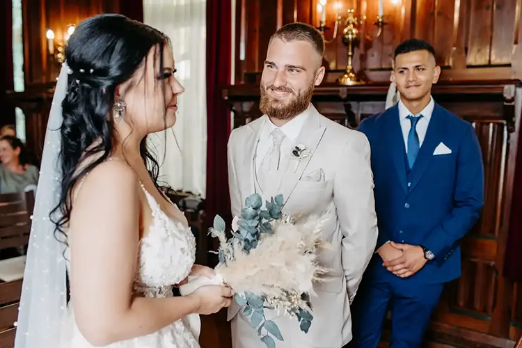 In einem mit Kronleuchtern geschmückten Holzraum steht eine Braut in einem weißen Kleid mit ihrem Brautstrauß und blickt dem Bräutigam in seinem hellen Anzug entgegen. Hinter ihnen steht ein Mann in einem blauen Anzug und beobachtet still diese magische Hochzeit, die von Melanie Kunert Fotografie wunderschön eingefangen wurde.