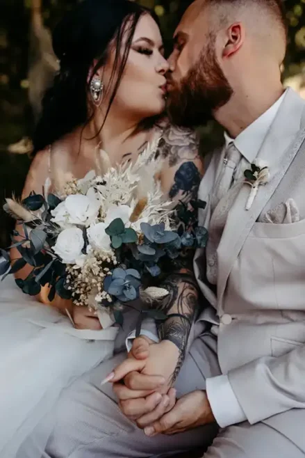 Das Paar küsst sich zärtlich während seiner Hochzeit in Tharandt. Die Braut sitzt eng beieinander und hat ihre Hände liebevoll ineinander verschlungen. In einem ärmellosen Kleid, das ihren tätowierten Arm freigibt, hält sie einen Strauß weißer und blauer Blumen in der Hand. Neben ihr steht der Bräutigam in seinem beigen Anzug und seiner Krawatte und sieht dabei elegant aus.