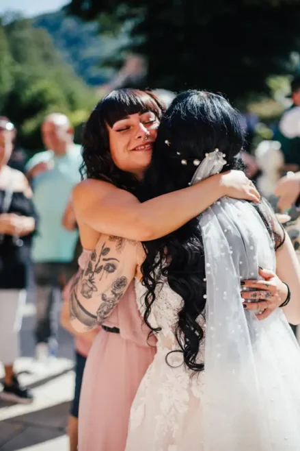 Eine Frau in einem hellrosa Kleid und mit tätowierten Armen umarmt freudig eine Braut mit langem, schwarzem, perlengeschmücktem Haar während einer Hochzeit in Tharandt. Sie stehen im Freien, umgeben von den anderen Gästen an diesem sonnigen Tag.