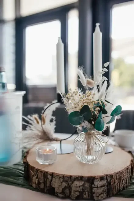 Ein dekorativer Tisch für eine Hochzeit in Tharandt besteht aus einer Holzplatte mit einem kleinen Strauß Trockenblumen in einer Glasvase, zwei weißen Stabkerzen in Kerzenhaltern und einem brennenden Teelicht. Der sanft verschwommene Hintergrund wird durch das Fensterlicht wunderschön beleuchtet.