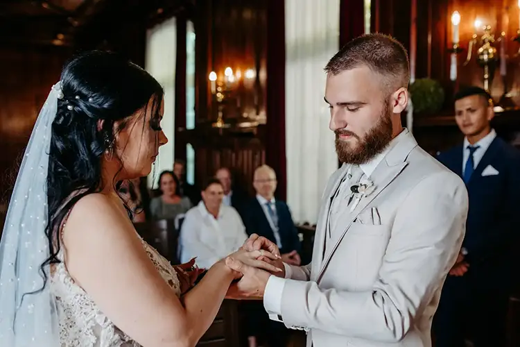 In einem eleganten, schwach beleuchteten Raum tauschen Braut und Bräutigam während ihrer Hochzeit in Tharandt die Ringe. Die Braut strahlt in ihrem weißen Kleid und Schleier, während der Bräutigam in einem hellen Anzug elegant aussieht. Melanie Kunert Fotografie fängt die Szene ein, während die Gäste aufmerksam von ihren Plätzen aus zuschauen.