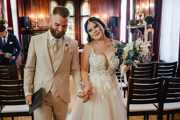 Ein Paar in Hochzeitskleidung schreitet anmutig den Gang entlang. Die Braut, einen Blumenstrauß in der Hand, beeindruckt in ihrem tief ausgeschnittenen Kleid. Der Bräutigam im hellen Anzug hält eine Broschüre in der Hand. Holzstühle und Buntglas bilden die Kulisse, wunderschön eingefangen von Hochzeit in Tharandt-Melanie Kunert Fotografie.