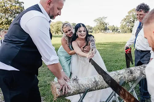Eine Braut im weißen Kleid sägt fröhlich einen Baumstamm. Unterstützt wird sie dabei von einer Brautjungfer und zwei Männern in festlicher Kleidung. Die Gruppe ist auf einer Wiese im Freien und voll und ganz dieser bezaubernden Tradition gewidmet.