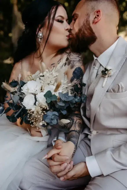 Das Paar küsst sich zärtlich während seiner Hochzeit in Tharandt. Die Braut sitzt eng beieinander und hat ihre Hände liebevoll ineinander verschlungen. In einem ärmellosen Kleid, das ihren tätowierten Arm freigibt, hält sie einen Strauß weißer und blauer Blumen in der Hand. Neben ihr steht der Bräutigam in seinem beigen Anzug und seiner Krawatte und sieht dabei elegant aus.