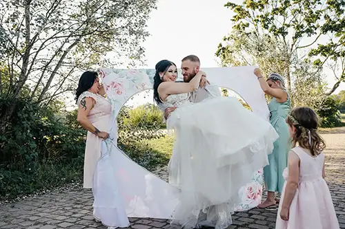 Ein Bräutigam trägt die Braut verspielt in seinen Armen, während sie in einem floralen Bilderrahmen im Freien bei ihrer Hochzeit in Tharandt posieren. Drei Brautjungfern assistieren, während ein junges Blumenmädchen in einem weißen Kleid zusieht. Im Hintergrund sind Bäume und ein sonnenbeschienener Weg zu sehen.