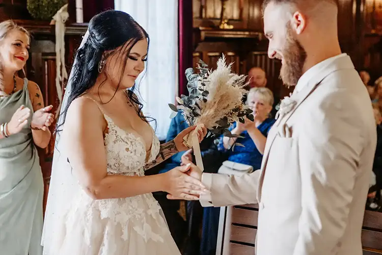 Bei ihrer Hochzeit in Tharandt, festgehalten von Melanie Kunert Fotografie, tauschen Braut und Bräutigam die Ringe. Die Braut, in einem Spitzenkleid, strahlt, als sie ihrem Bräutigam den Ring an den Finger steckt. Er steht in einem hellen Anzug da und lächelt zurück, während die Gäste diesen lieblichen Moment miterleben.
