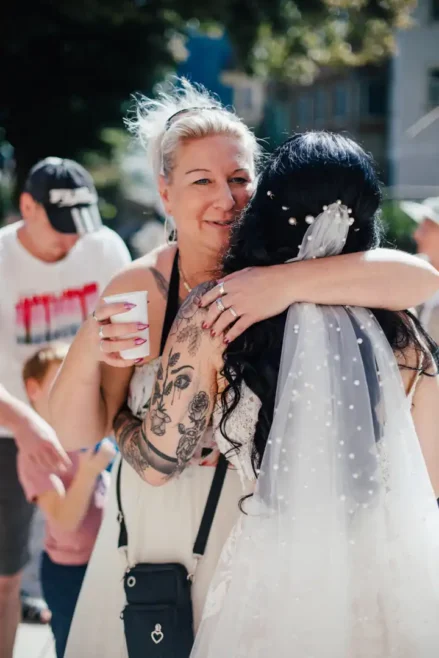Eine Frau mit Tattoos und einer schwarzen Tasche umarmt eine Braut mit langen schwarzen Haaren und einem perlenbesetzten Schleier bei einer Hochzeit im Freien in Tharandt. Die Frau hält eine Tasse, während Kinder und Erwachsene im Hintergrund den sonnigen Tag genießen.