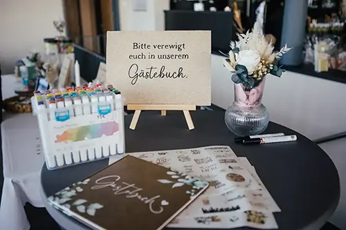 Ein runder Tisch mit bunten Stiften, einem Gästebuch und dekorativen Aufklebern steht elegant da. Daneben steht auf einer Staffelei ein Schild mit der Aufschrift „Bitte verewigt euch in unserem Gästebuch“, neben einer Blumenvase – die perfekte Kulisse für eine bezaubernde Hochzeit in Tharandt.