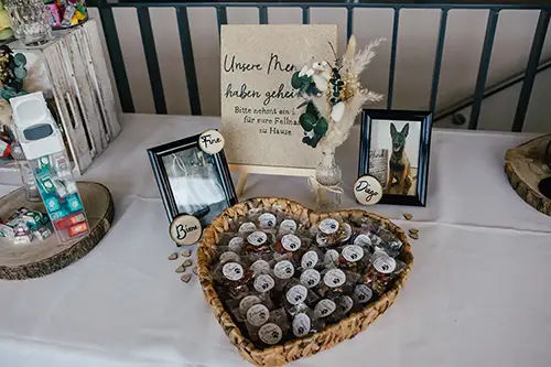 Auf einem Tisch steht ein herzförmiger Korb mit kleinen Gläsern, eingerahmt von zwei Fotos. Ein beiges Schild mit deutschem Text deutet dezent auf eine Hochzeit in Tharandt hin. Die Dekoration besteht aus charmanten Holzschildern, einer Vase mit Laub und verstreuten Kieselsteinen für eine elegante Note.