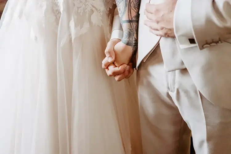 Bei ihrer Hochzeit in Tharandt hält ein Paar im Hochzeitskleid Händchen. Das weiße Kleid der Braut ist mit zarten Spitzendetails verziert und harmoniert perfekt mit dem hellbeigen Anzug des Bräutigams. Ein dezentes Tattoo ziert ihren Unterarm und verleiht ihrem besonderen Tag eine persönliche Note.