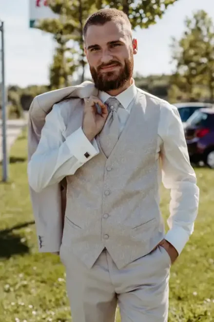 Ein bärtiger Mann steht im Freien bei einer Hochzeit in Tharandt. Er trägt eine beige Weste und ein passendes Jackett über der Schulter. Eine Hand steckt in der Tasche und lächelt in die Kamera. Im sonnenbeschienenen Hintergrund sind Bäume und ein Auto zu sehen.