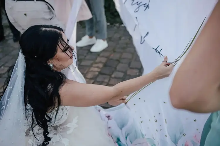 Eine Braut kniet auf Kopfsteinpflaster inmitten einer Hochzeit in Tharandt. Ihr Schleier und ihr Kleid wehen, während sie mit einer Schere ein großes Banner zerschneidet. Umgeben von teilweise sichtbaren Gästen fängt die Szene im Freien die Essenz der Feier ein.