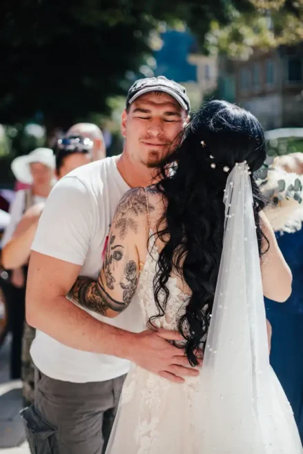 Eine Braut mit langem, dunklem Haar und Perlenschleier umarmt einen Mann in weißem T-Shirt und Mütze unter dem sonnigen Himmel von Hochzeit in Tharandt. Im unscharfen Hintergrund versammeln sich Menschen, während der tätowierte Arm des Mannes den Charme des Augenblicks verstärkt.
