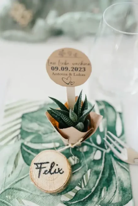 Nahaufnahme einer Hochzeitstafel . Darin zu sehen ist eine kleine Sukkulente im Topf mit einem Holzschild mit der Aufschrift „09.09.2023 Antonia & Lukas“. Daneben liegt eine hölzerne Tischkarte mit der Aufschrift „Felix“ auf einer Stoffserviette mit Blattmuster, dazu eine Gabel und ein Glas.