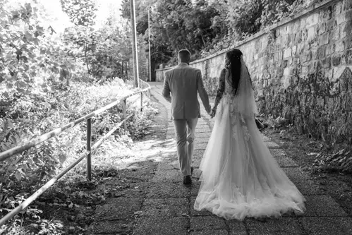 Braut und Bräutigam gehen Hand in Hand einen sonnenbeschienenen Weg entlang, gesäumt von Bäumen und einer Steinmauer. Ein wunderschönes Foto von Melanie Kunert Fotografie. Das lange, fließende Brautkleid und der Anzug des Bräutigams unterstreichen diese ruhige Hochzeit in Tharandt und schaffen eine wahrhaft romantische Szene.