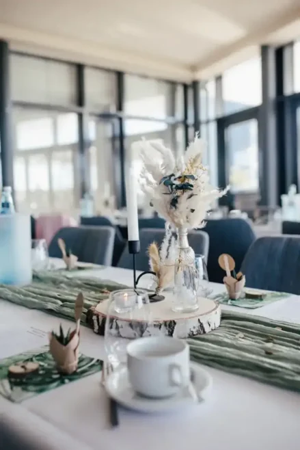 Ein dekorativer Tisch für eine Hochzeit besteht aus einer Holzplatte mit einem kleinen Strauß Trockenblumen in einer Glasvase, zwei weißen Stabkerzen in Kerzenhaltern und einem brennenden Teelicht. Der sanft verschwommene Hintergrund wird durch das Fensterlicht wunderschön beleuchtet.