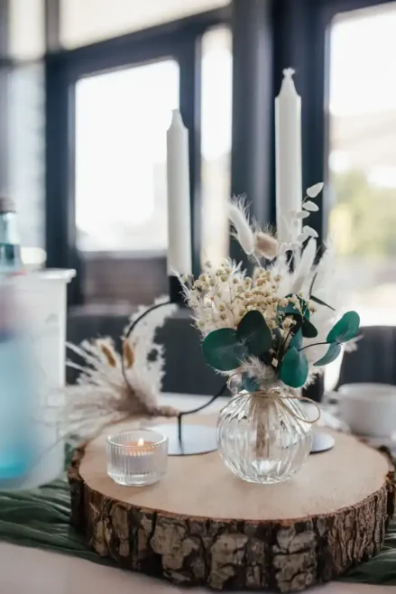 Ein dekorativer Tisch für eine Hochzeit besteht aus einer Holzplatte mit einem kleinen Strauß Trockenblumen in einer Glasvase, zwei weißen Stabkerzen in Kerzenhaltern und einem brennenden Teelicht. Der sanft verschwommene Hintergrund wird durch das Fensterlicht wunderschön beleuchtet.