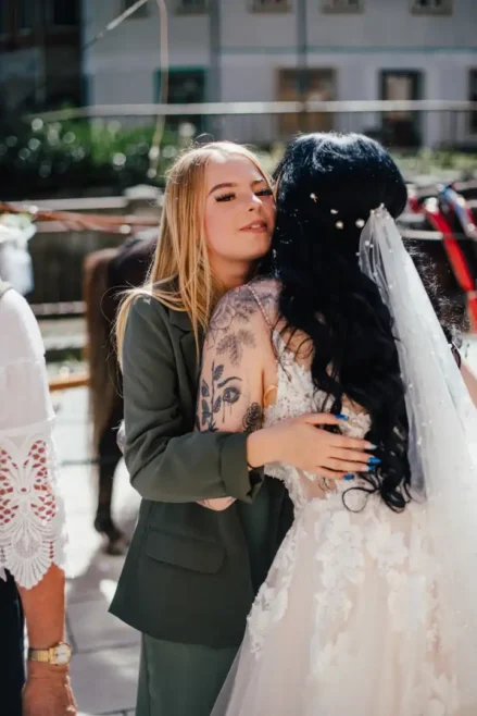 Eine Braut in weißem Brautkleid und Schleier wird von einer Frau mit langen blonden Haaren in einem grünen Anzug umarmt. Die Braut trägt ein Blumentattoo auf dem Arm. Die beiden stehen draußen in einer sonnigen Umgebung mit Gebäuden im Hintergrund. Ein wunderschönes Foto von Melanie Kunert Fotografie bei Hochzeit in Tharandt.