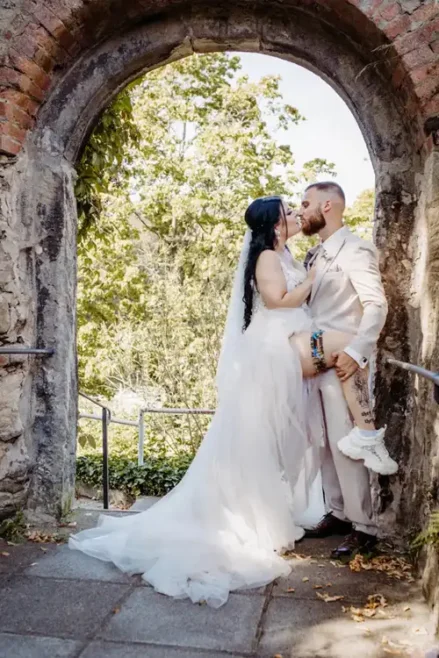 Braut und Bräutigam küssen sich unter einem steinernen Torbogen, wunderschön eingefangen von Melanie Kunert Fotografie. Die Braut in ihrem weißen Kleid und Schleier hebt verspielt ein Bein, während der Bräutigam in einem hellen Anzug daneben steht. Grünes Laub aus Tharandt bildet eine üppige Kulisse für ihren Hochzeitsmoment.