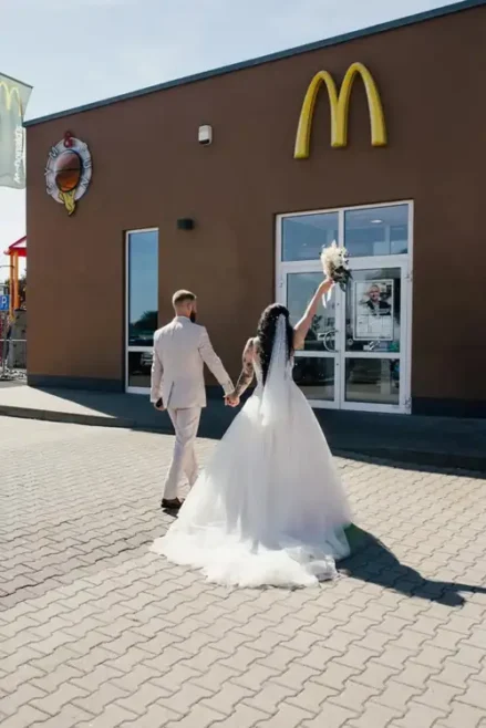 Braut und Bräutigam feiern ihre Hochzeit und gehen Hand in Hand auf einen McDonald's-Laden zu. Die Braut hält ihren Brautstrauß hoch, als sie sich dem Eingang nähern, über ihnen leuchtet das ikonische McDonald's-Logo.