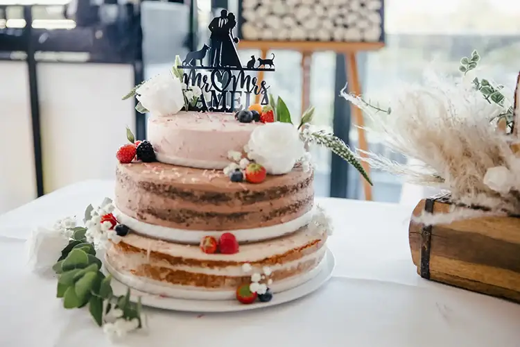 Eine dreistöckige, halbnackte Hochzeitstorte, inspiriert von einer Hochzeit in Tharandt, ist mit frischen Beeren, Grünpflanzen und weißen Blumen geschmückt. Gekrönt wird die Torte von einem „Mr. & Mrs. Cake“-Paar und einer Haustierfigur. Elegant ruht sie auf einer weißen Tischdecke.