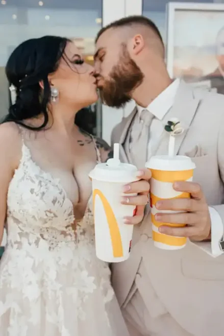 Ein Paar küsst sich und hält große Fast-Food-Getränke in der Hand. Die Braut glänzt in ihrem Spitzenbrautkleid, während der Bräutigam in seinem beigen Anzug mit Krawatte elegant aussieht und diesen ungezwungenen Moment draußen genießt.