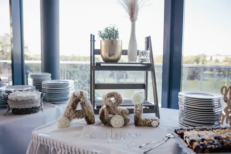 Tisch mit gestapelten Tellern, einem Desserttablett und den Initialen „A & L“ aus Schnur und Blumen. In Anlehnung an eine Hochzeit in Tharandt präsentiert er eine kleine Pflanze und eine Vase auf einem Holzregal. Große Fenster bieten einen malerischen Ausblick.