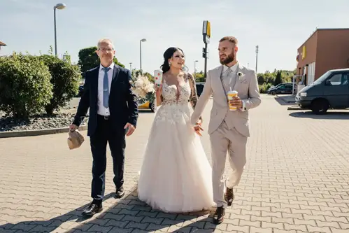 Eine Braut im weißen Kleid und ein Bräutigam im beigen Anzug feiern ihre Hochzeit in Tharandt Hand in Hand mit einem Mann im Anzug und einer Tasche in der Hand. Der Bräutigam genießt einen Drink, während sie über den Parkplatz schlendern, umgeben von Gebäuden und Grünflächen.