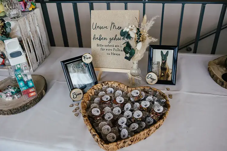 Auf einem Tisch steht ein herzförmiger Korb mit kleinen Gläsern, eingerahmt von zwei Fotos. Ein beiges Schild mit deutschem Text deutet dezent auf eine Hochzeit hin. Die Dekoration besteht aus charmanten Holzschildern, einer Vase mit Laub und verstreuten Kieselsteinen für eine elegante Note.