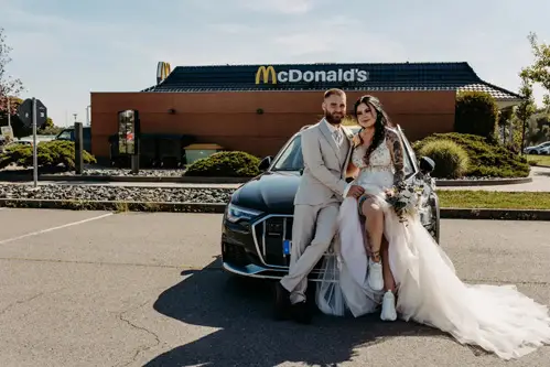 Ein Paar im Hochzeitskleid sitzt auf der Motorhaube eines Autos auf einem McDonald's-Parkplatz und feiert seine Hochzeit in Tharandt. Die Braut reckt aufgeregt ihren Brautstrauß in die Höhe. Sie trägt ein wallendes weißes Kleid und Turnschuhe, während der Bräutigam in seinem hellen Anzug lächelt. Im Hintergrund ist das McDonald's-Schild zu sehen.