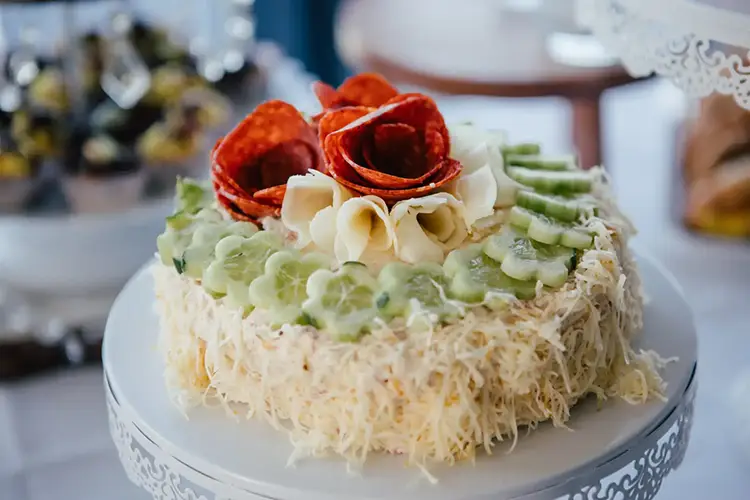 Eine dekorative Torte ziert die Hochzeit in Tharandt. Die Oberseite ist mit Gurkenscheiben verziert, die ein elegantes Muster bilden. In der Mitte schmiegen sich Rosetten aus Peperoni und Käse aneinander. Die Torte ruht auf einem kunstvollen weißen Ständer und ist in sanftes Licht getaucht.
