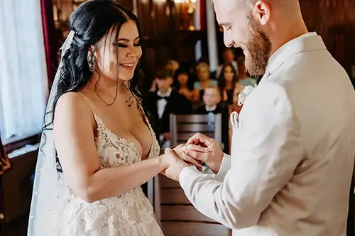 Bei ihrer Hochzeit in Tharandt, festgehalten von Melanie Kunert Fotografie, tauschen Braut und Bräutigam die Ringe. Die Braut, in einem Spitzenkleid, strahlt, als sie ihrem Bräutigam den Ring an den Finger steckt. Er steht in einem hellen Anzug da und lächelt zurück, während die Gäste diesen lieblichen Moment miterleben.