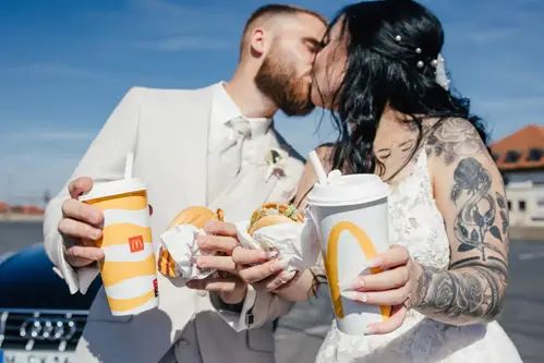 Bei einer malerischen Hochzeitt küsst sich ein frisch vermähltes Paar im Hochzeitskleid und hält McDonald's-Getränke und Burger in den Händen. Der Bräutigam trägt einen hellen Anzug, während die tätowierte Braut in Weiß umwerfend aussieht. Sie stehen draußen neben einem Auto unter dem strahlend blauen Himmel.