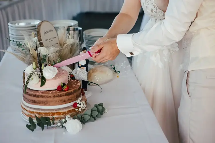 Ein Paar schneidet bei seiner Hochzeit eine dreistöckige Torte an, die mit Blumen und Erdbeeren geschmückt ist. Die Torte steht wunderschön auf einem Tisch mit weißer Tischdecke, im Hintergrund zieren gestapelte Teller die Torte.