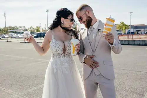 Eine Braut im weißen Spitzenkleid und ein Bräutigam im beigen Anzug lachen und halten draußen auf einem Tharandter Parkplatz Fastfood in der Hand. Fröhlich, mit Sonnenbrille und vom Wind zerzaustem Haar verkörpern sie den Geist ihres Hochzeitsabenteuers.