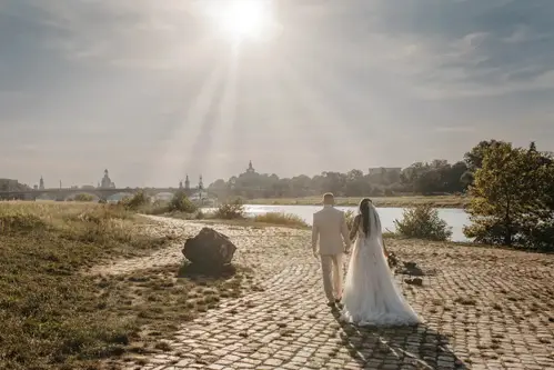 Ein Paar in Hochzeitskleidung hält Händchen auf einem Kopfsteinpflasterweg an einem Fluss. Im Hintergrund ist die Silhouette von Dresden zu sehen. Der Himmel ist klar und sonnig und wirft ein sanftes Licht auf die Hochzeitsszene.