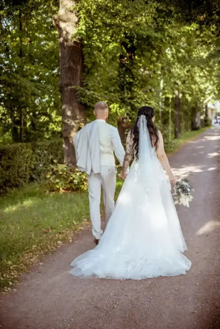 Braut und Bräutigam gehen Hand in Hand einen von Bäumen gesäumten Weg in Tharandt entlang. Die Braut trägt ein fließendes weißes Kleid mit einem langen Schleier, während der Bräutigam, der ihre Hochzeit feiert, einen hellen Anzug trägt und sein Jackett über der Schulter trägt. Sonnenlicht fällt durch die Blätter.