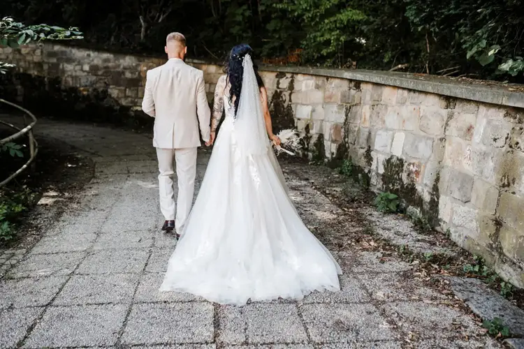 Braut und Bräutigam gehen Hand in Hand einen sonnenbeschienenen Weg entlang, gesäumt von Bäumen und einer Steinmauer. Ein wunderschönes Foto von Melanie Kunert Fotografie. Das lange, fließende Brautkleid und der Anzug des Bräutigams unterstreichen diese ruhige Hochzeit in Tharandt und schaffen eine wahrhaft romantische Szene.