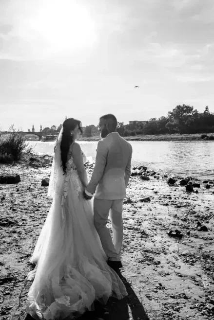 Braut und Bräutigam stehen Händchen haltend an einem felsigen Ufer unter strahlendem Himmel während ihrer Hochzeit. Die Braut trägt ein wallendes Kleid, der Bräutigam einen hellen Anzug. Im Hintergrund sind Bäume und ein Fluss zu sehen.