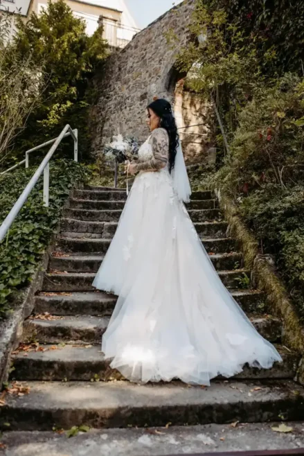 Bei der zauberhaften Hochzeit in Tharandt schreitet eine Braut in ihrem weißen Kleid über die Steintreppe. Mit ihrem langen, dunklen Haar, das ihr herabfällt, hält sie einen Blumenstrauß in den Händen. Die rustikale Szene wird von einer Steinmauer und üppigem grünem Laub eingerahmt, wunderschön eingefangen von Melanie Kunert Fotografie.